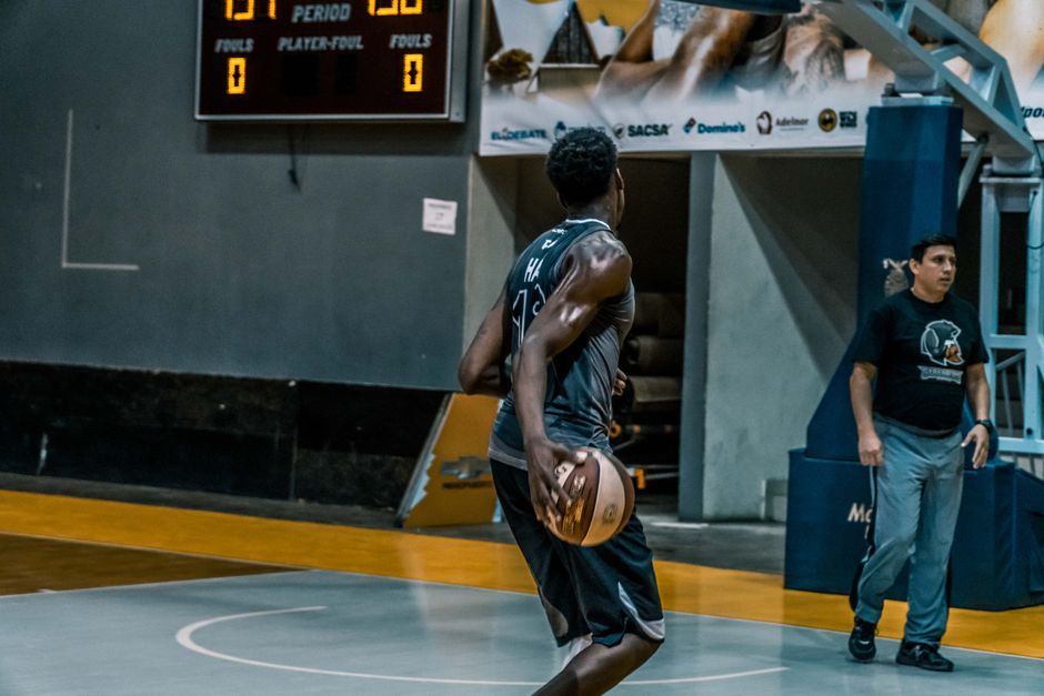 Basketball player performing on an indoor court with scoreboard in background, highlighting athleticism and teamwork.